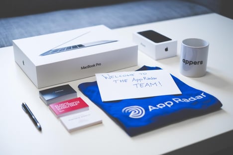 Items making up a new employee's welcome package are neatly arranged and seen as if the viewer was standing in front of it. It consists of a coffee mug, packaged iPhone and MacBook Pro, business cards, a pen, a t-shirt and a handwritten note that reads 'Welcome to the AppRadar Team'